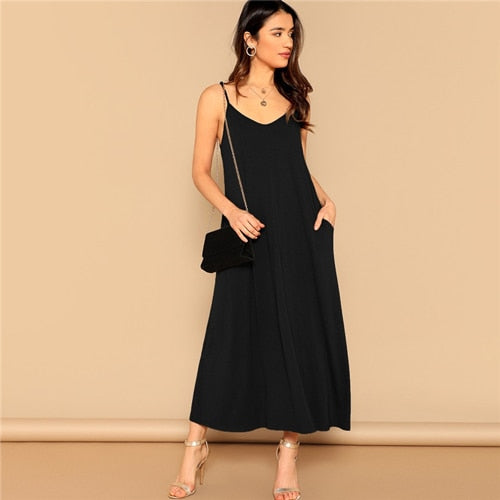 A model wearing a black sleeveless V-neck maxi dress with adjustable spaghetti straps and side pockets. The dress has a relaxed, flowy silhouette and is paired with gold heels and a black handbag.